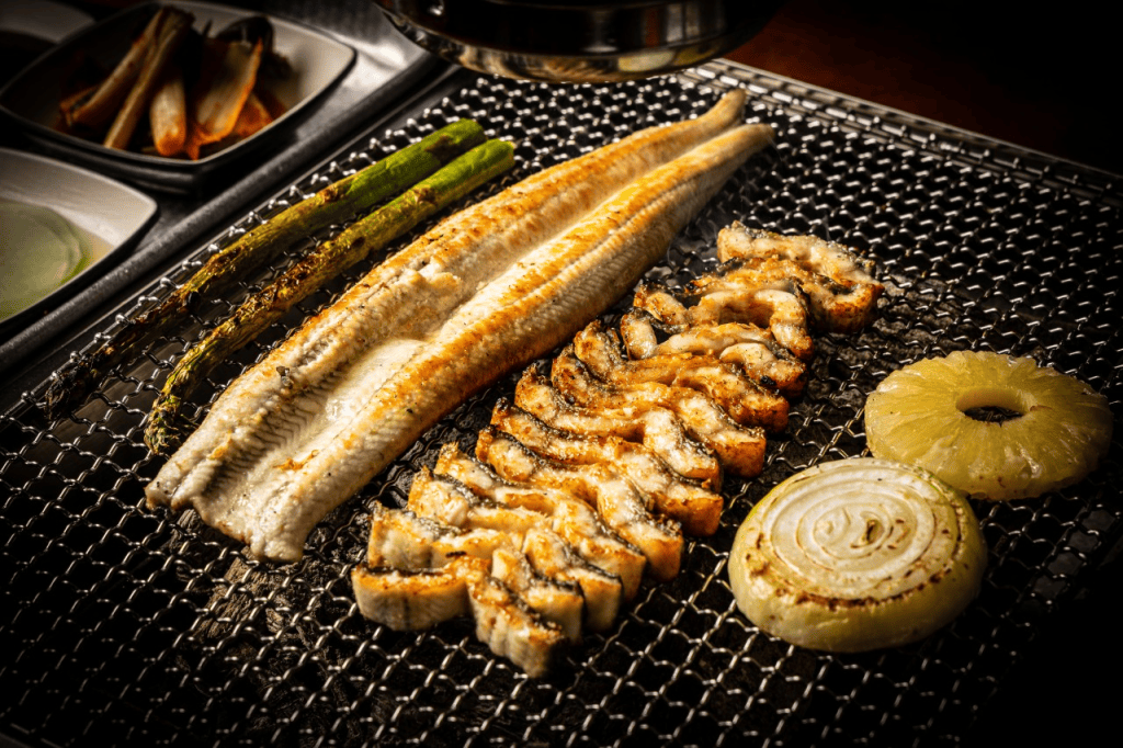 焼かれた魚と野菜がグリル上に並んでいる写真。白身魚、玉ねぎ、パイナップル、アスパラガスが含まれている。